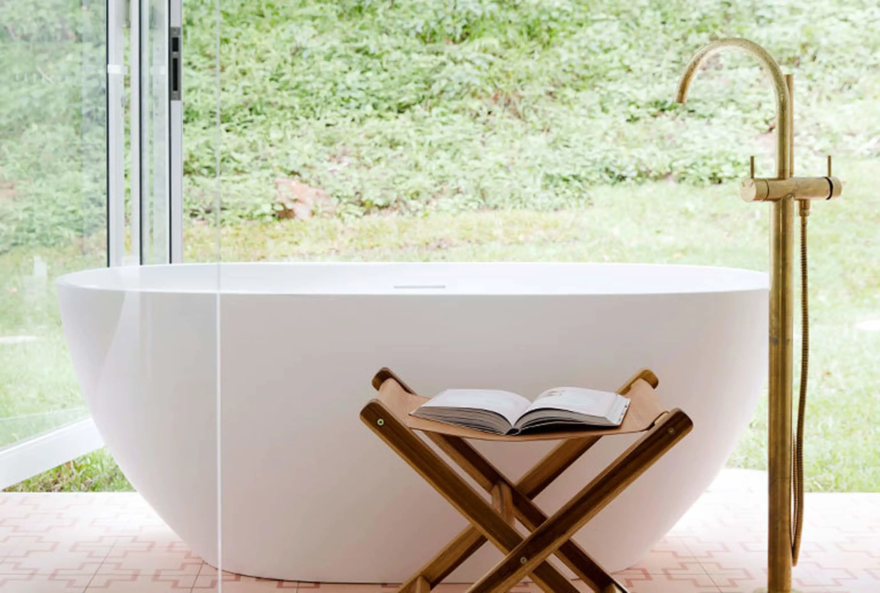 Modern white freestanding bathtub with brass fixtures, wooden stool holding open book, and large windows overlooking greenery.