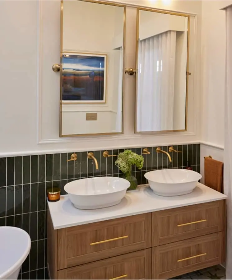 Modern bathroom with dual white vessel sinks, wooden vanity, green tile backsplash, and gold-framed mirrors.