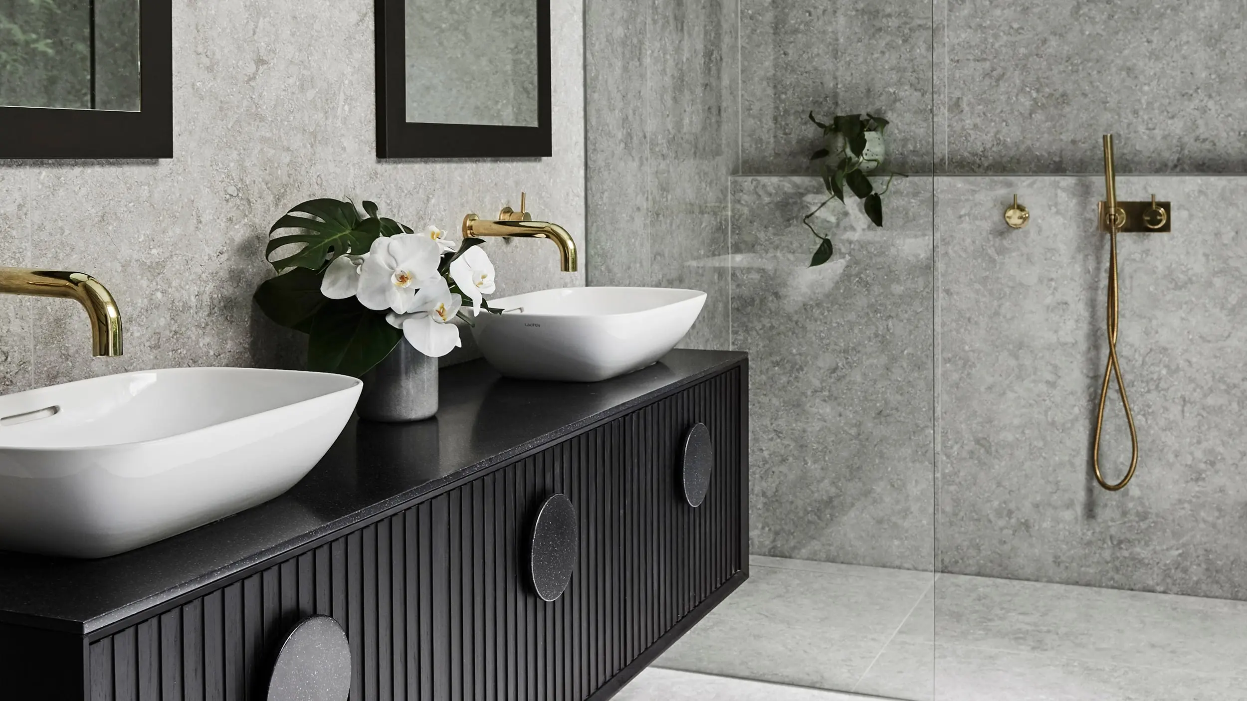 Modern bathroom with gray walls, black vanity, white vessel sinks, brass fixtures, and white orchids in a vase.