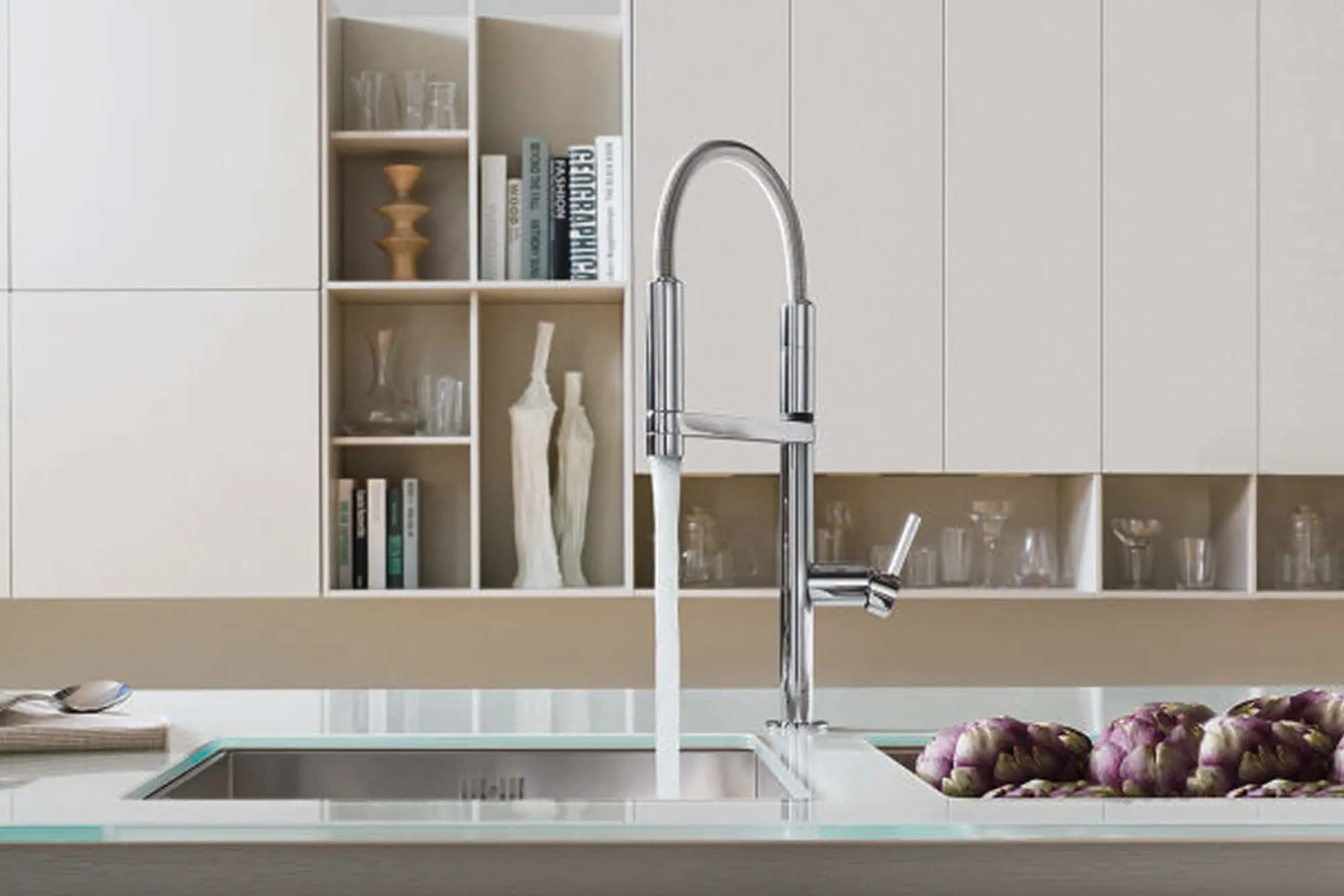 Modern kitchen sink with tall chrome faucet, glass countertop, artichokes, and built-in shelving with decorative items.