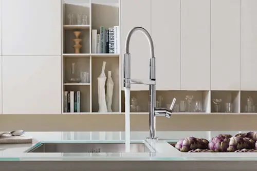 Modern kitchen sink with tall chrome faucet, glass countertop, artichokes, and built-in shelving with decorative items.