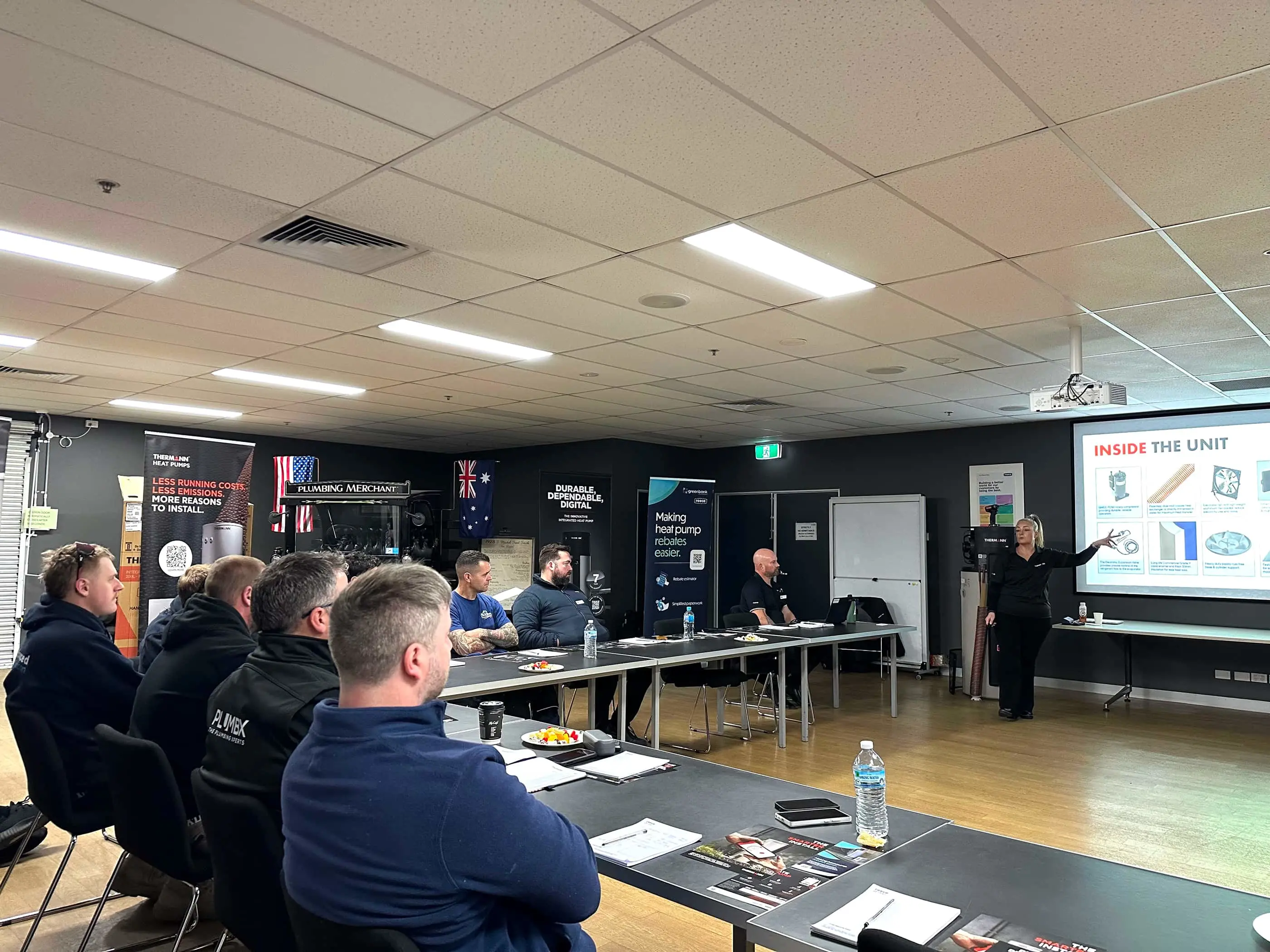 Plumbing training session with presenter explaining unit components to attendees seated at tables in a workshop classroom.