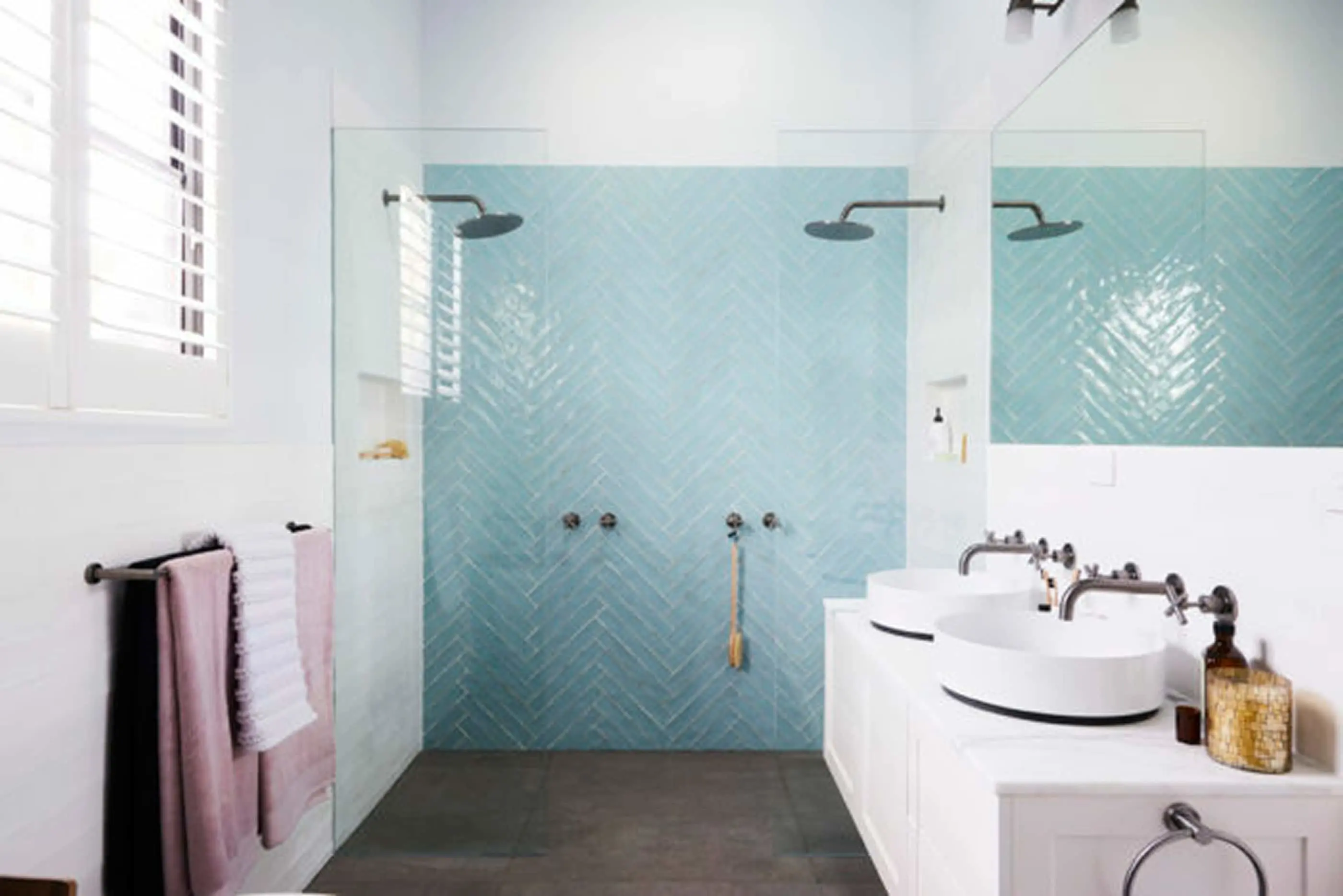 Modern bathroom with light blue herringbone tile shower, glass partition, white vessel sinks, and pink towels.
