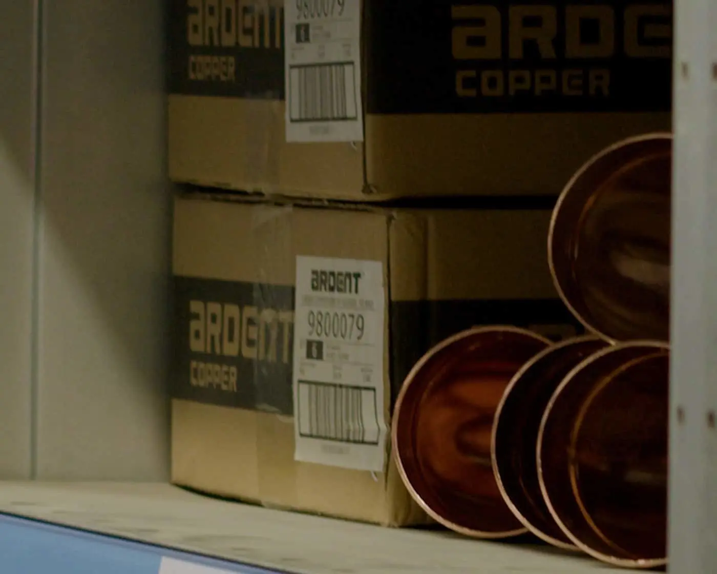 Stacked boxes of Ardent copper products with shipping labels next to copper pipes or tubes on a shelf.