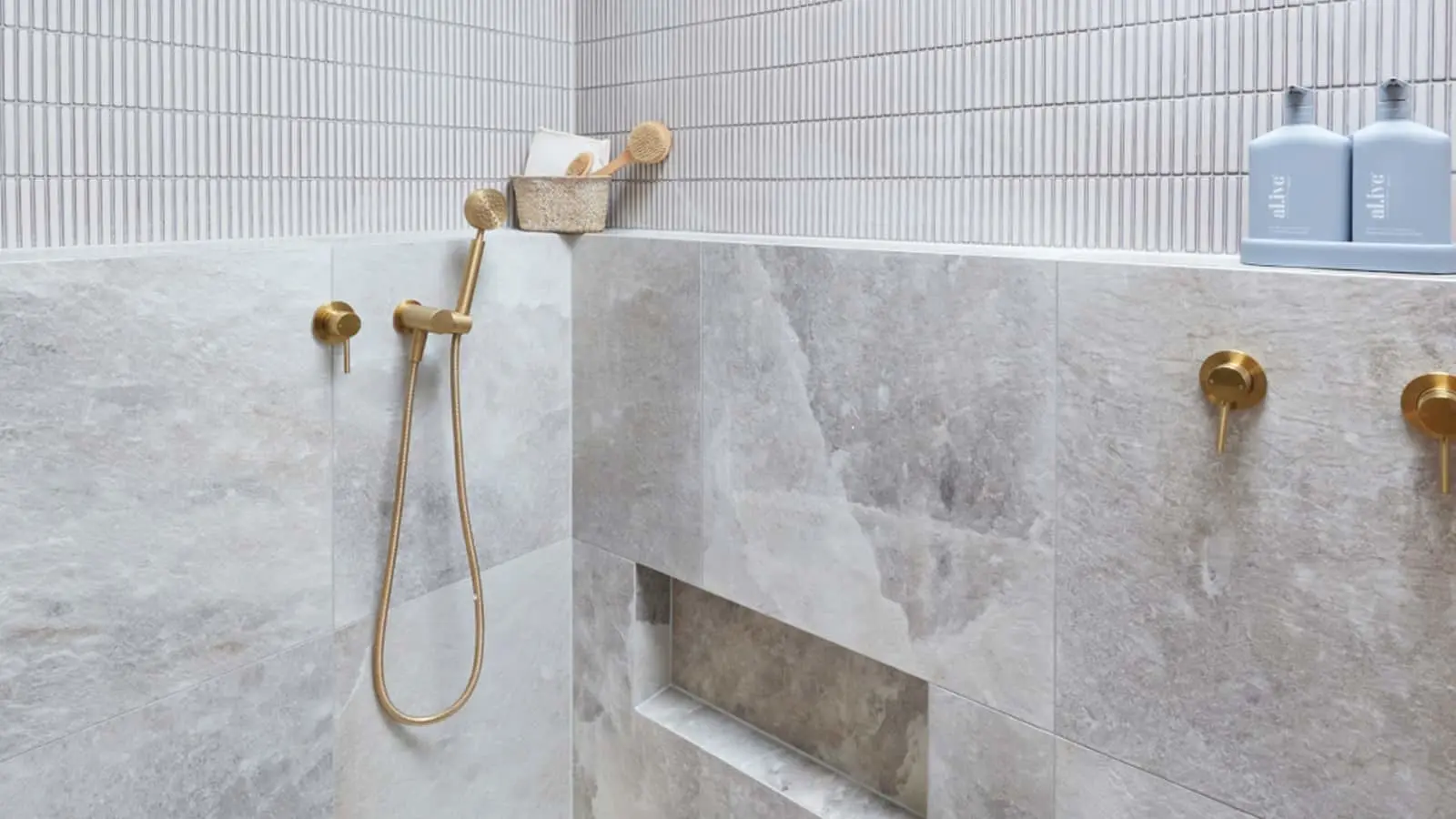 Modern bathroom with marble walls, brass fixtures, handheld shower, and bath products on a shelf.