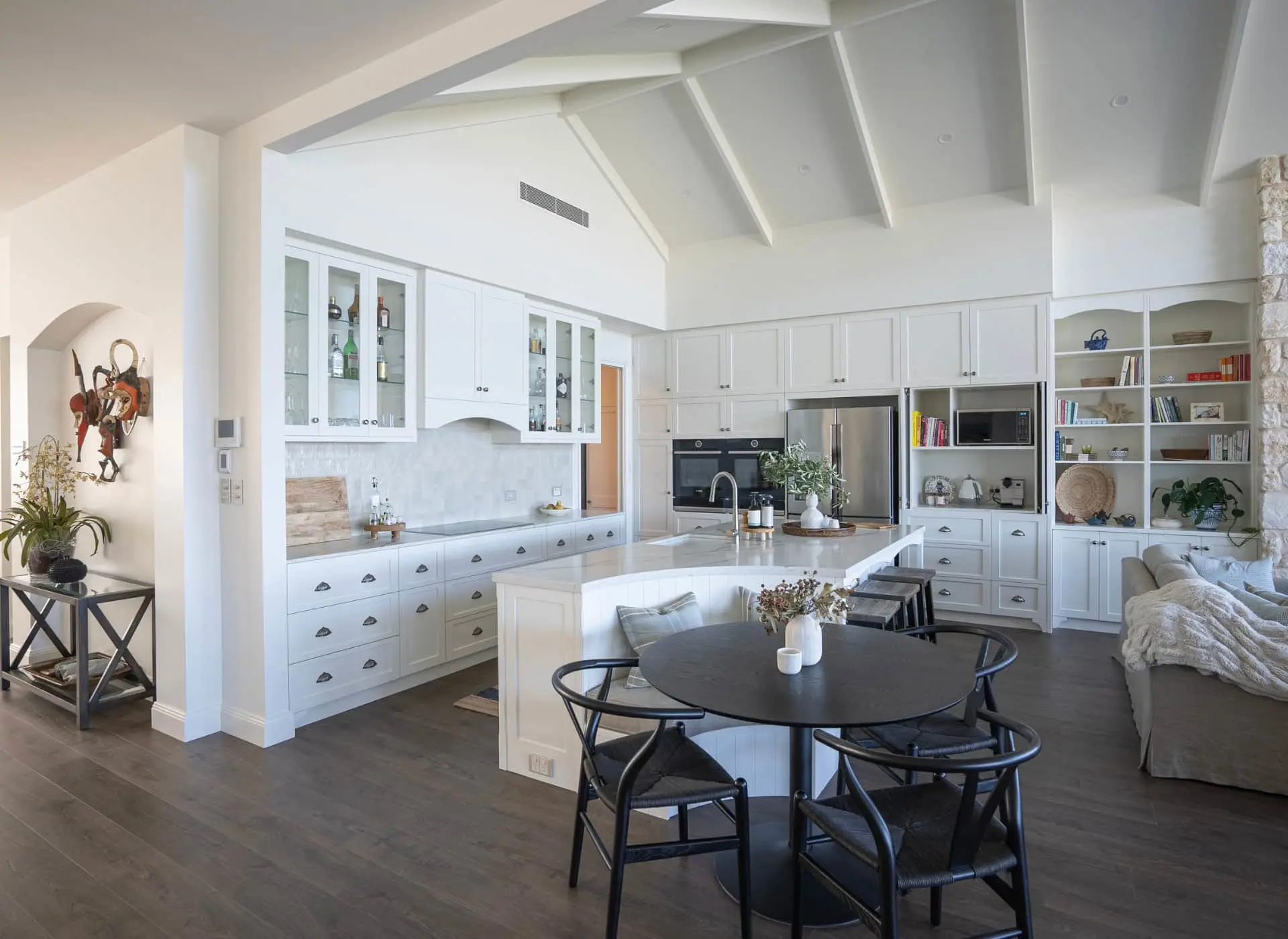 Modern white kitchen with vaulted ceiling, central island, black dining table, stainless steel appliances and built-in shelving.