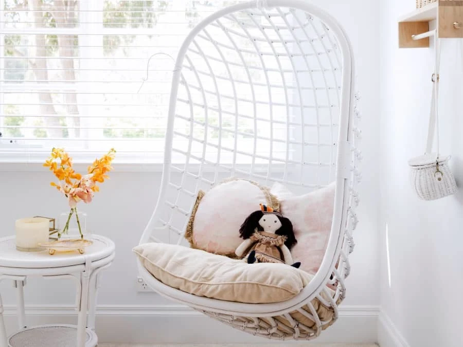 White hanging egg chair with beige cushions and a doll, next to a small table with orange flowers in a bright room.