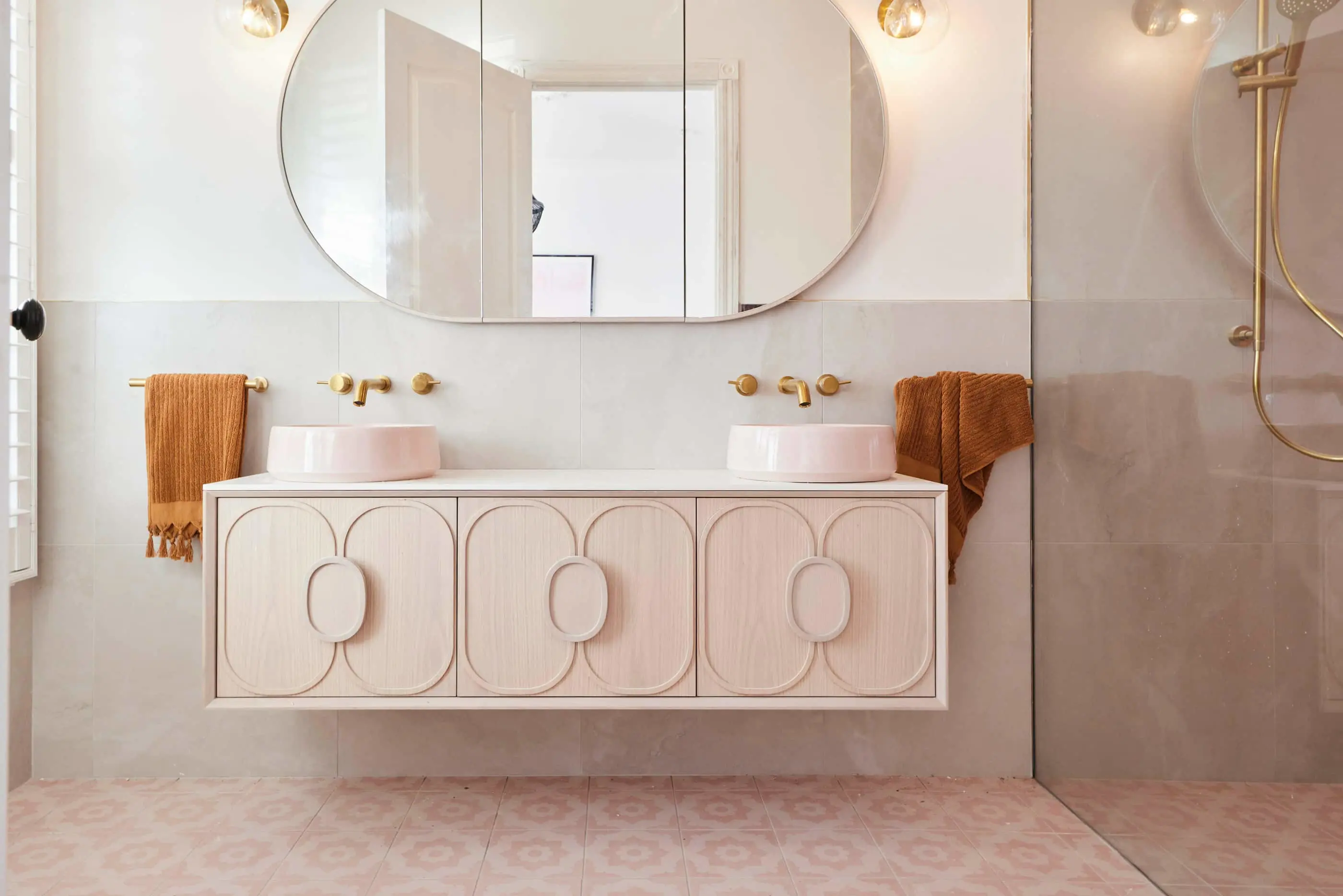 Modern bathroom with light wood vanity, two pink sinks, gold fixtures, oval mirror, and orange towels on pink tiled floor.