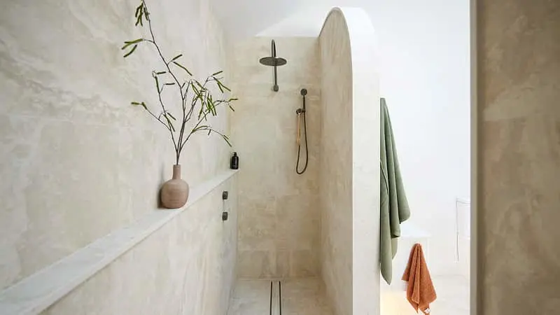 Modern minimalist bathroom with beige stone walls, walk-in shower, small plant in clay vase, and green and orange towels.