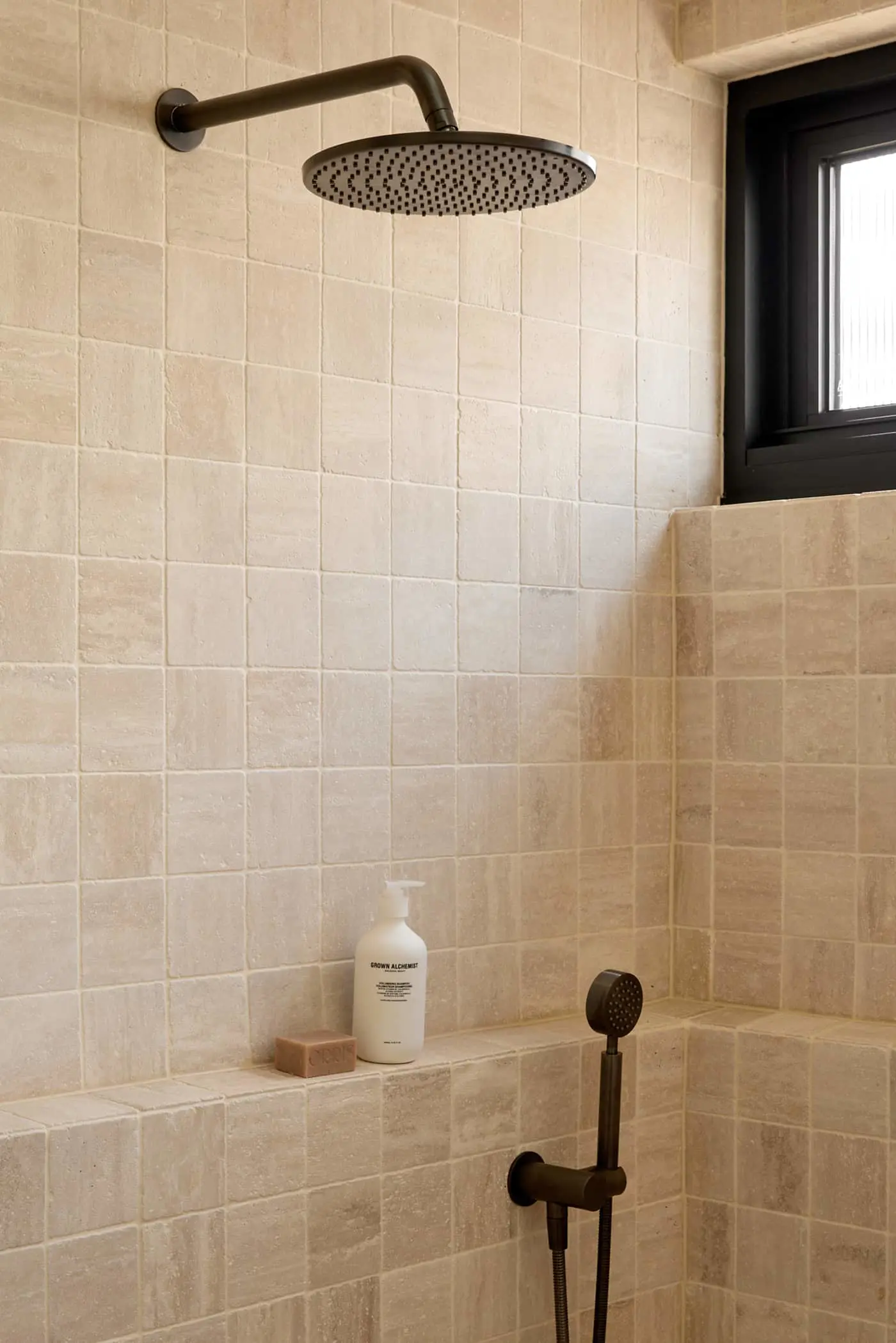 Modern bathroom shower with round black rainfall showerhead, handheld sprayer, and beige tiled walls with built-in shelf.