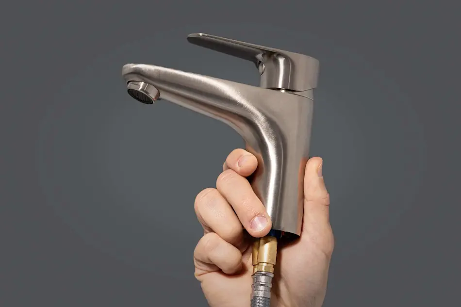 Hand holding a brushed stainless steel bathroom faucet with brass fitting against a gray background.