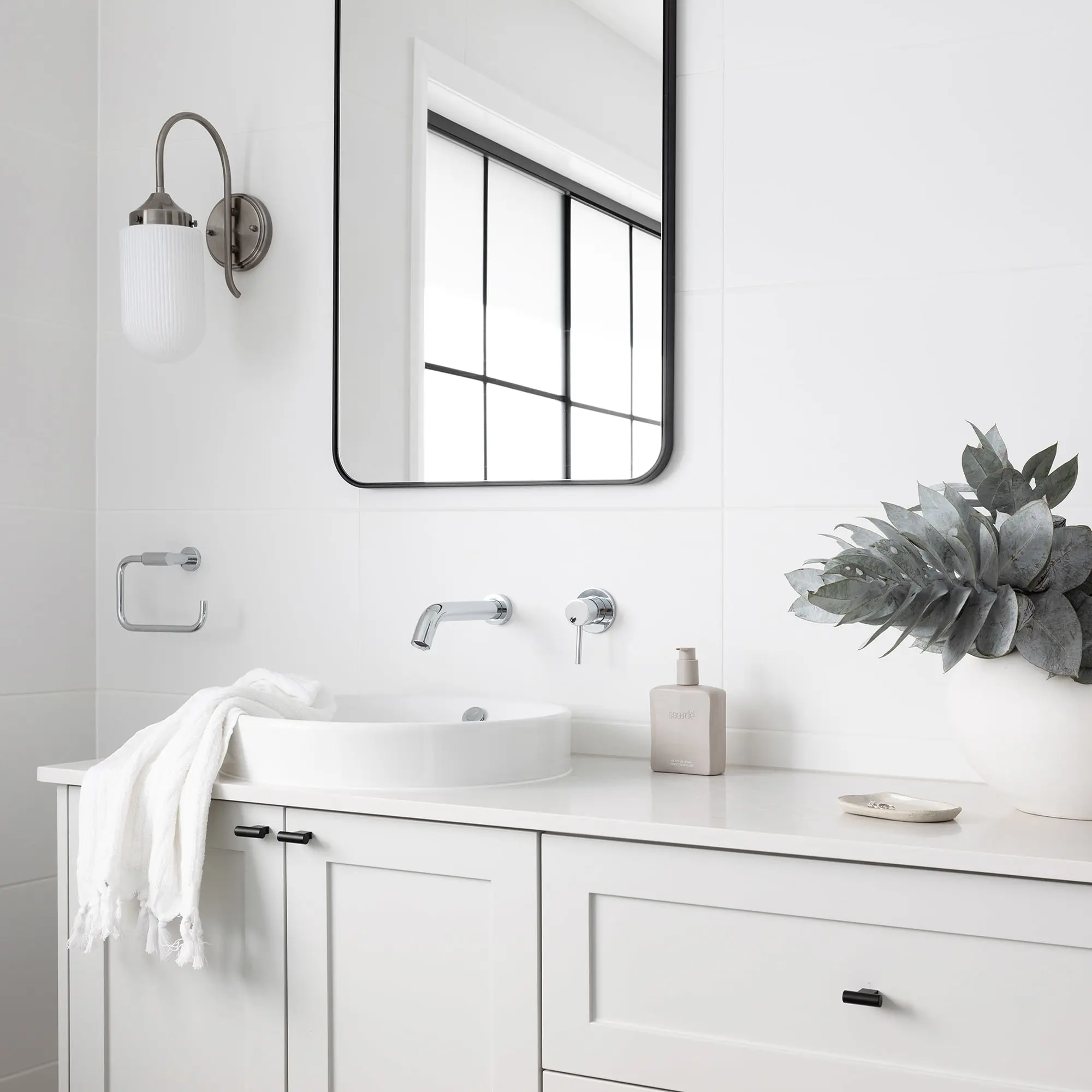 Modern white bathroom with vessel sink, wall-mounted faucet, black-framed mirror, sconce light, and decorative plant.