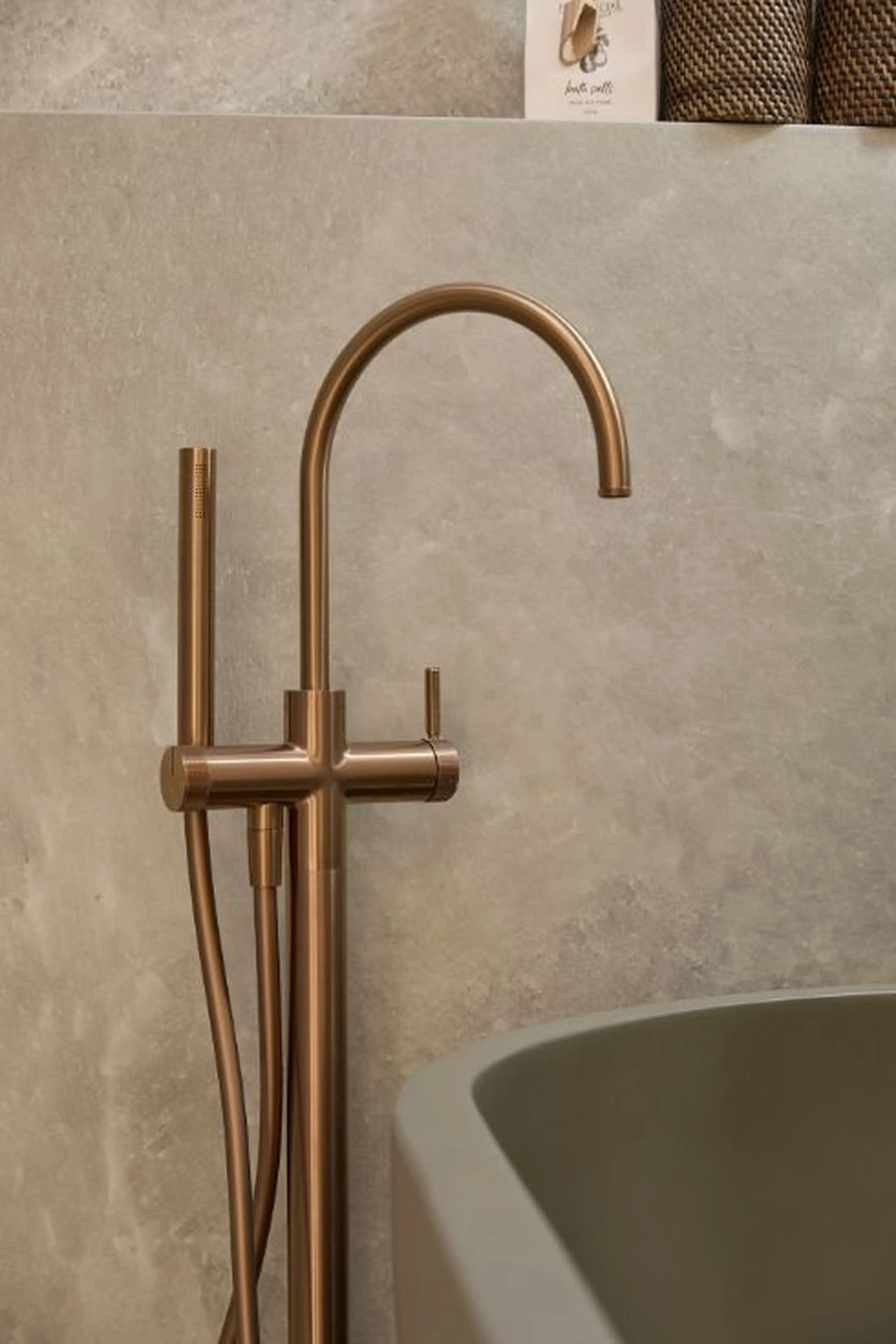 Modern brass freestanding bathtub faucet with curved spout against beige concrete wall, edge of tub visible.