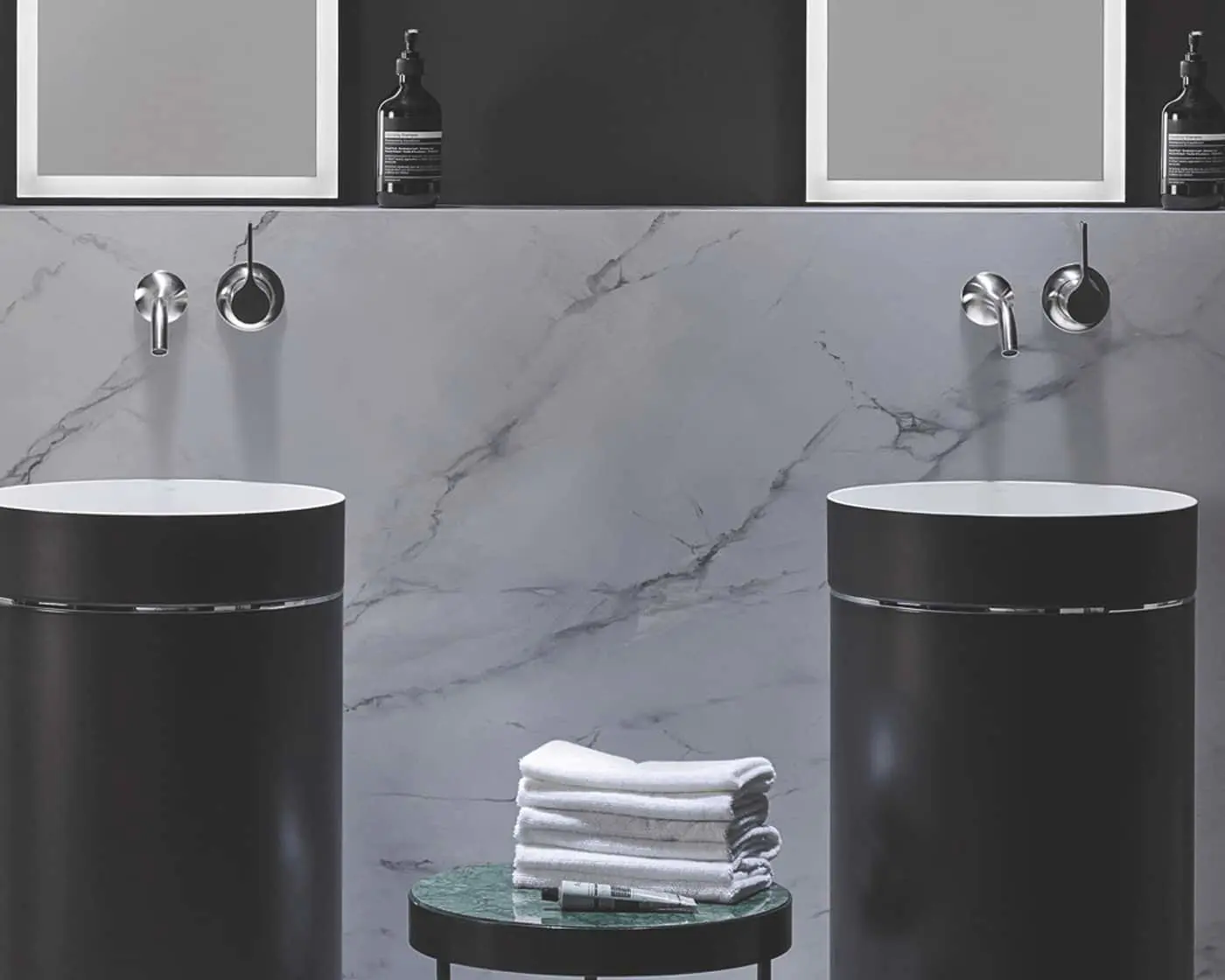 Modern bathroom with two black cylindrical sinks, marble wall, wall-mounted faucets, and folded white towels on a small table.