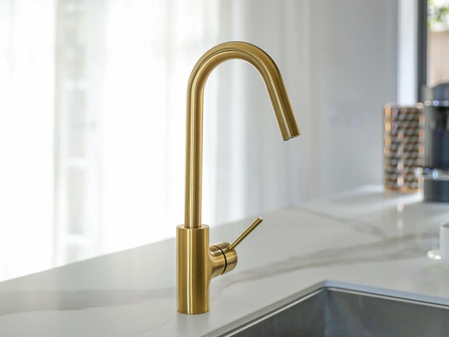 Modern brushed gold kitchen faucet with high curved spout on white marble countertop next to stainless steel sink.