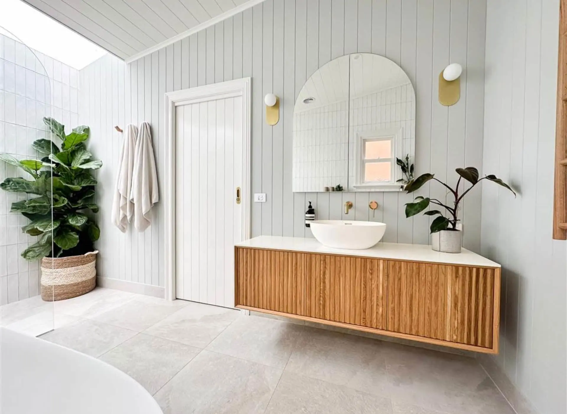 Modern bathroom with light gray walls, wooden vanity, round mirror, white vessel sink, and potted plants.