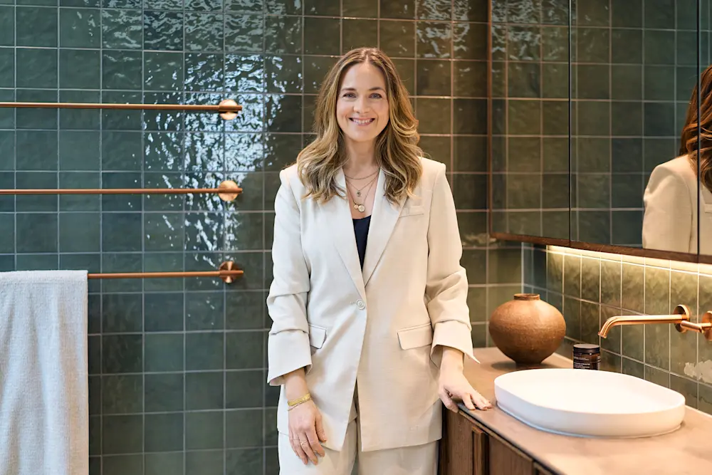 Person in cream blazer smiling in modern bathroom with green tile walls, copper fixtures, and wooden vanity.