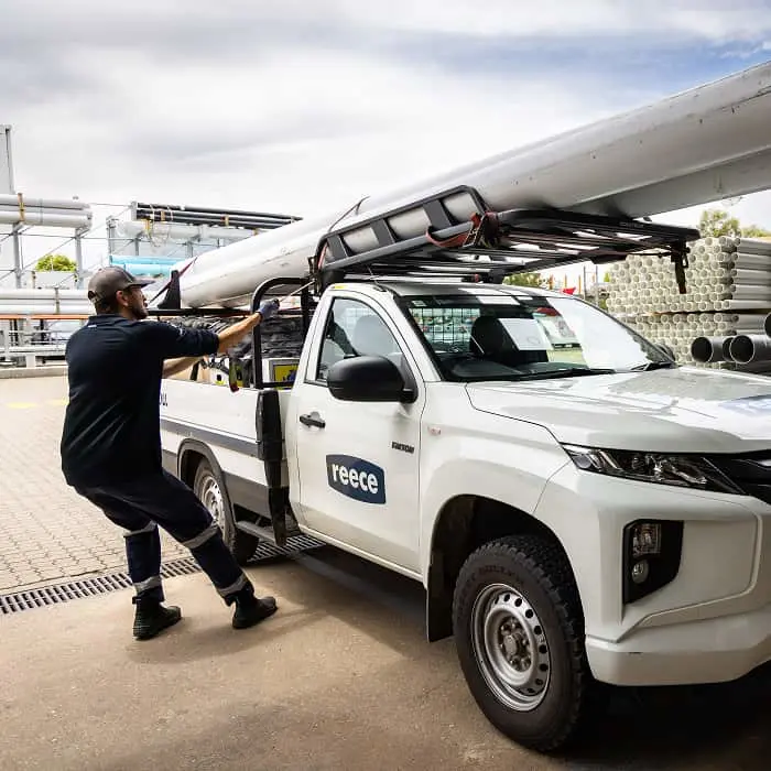 Person loading long pipes onto roof rack of white Reece pickup truck at industrial site under cloudy sky.