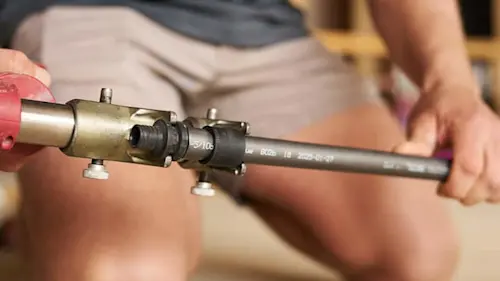 Person holding a pipe fitting tool with brass connector attached to a gray PEX pipe during installation.