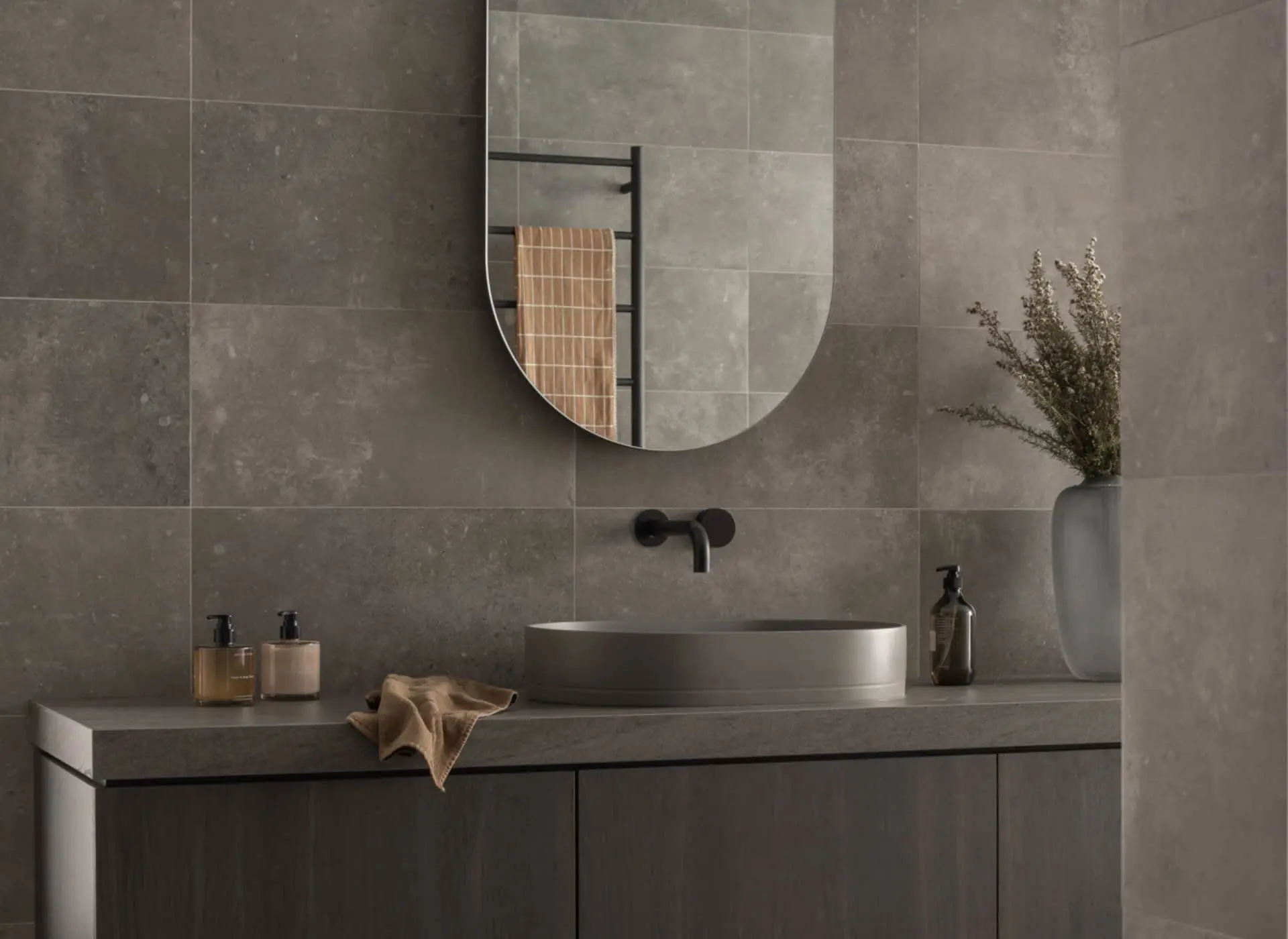 Modern bathroom with gray tile walls, oval mirror, vessel sink, black fixtures, and minimalist decor including dried plants.