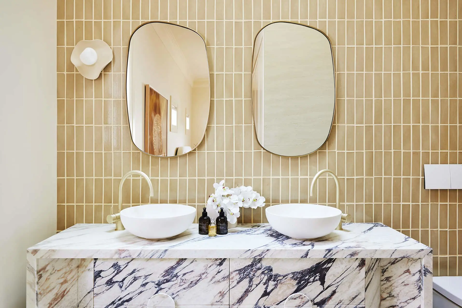 Modern bathroom with beige tiled wall, two oval mirrors, marble vanity with white vessel sinks, and white orchids.