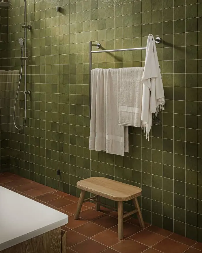 Bathroom with green tiled walls, white towels on metal bar, handheld shower, wooden bench, and terracotta floor tiles.