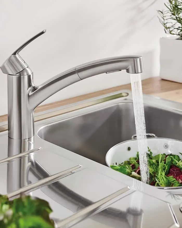 Stainless steel kitchen faucet running water into a colander of fresh greens over a sink with wooden countertop.