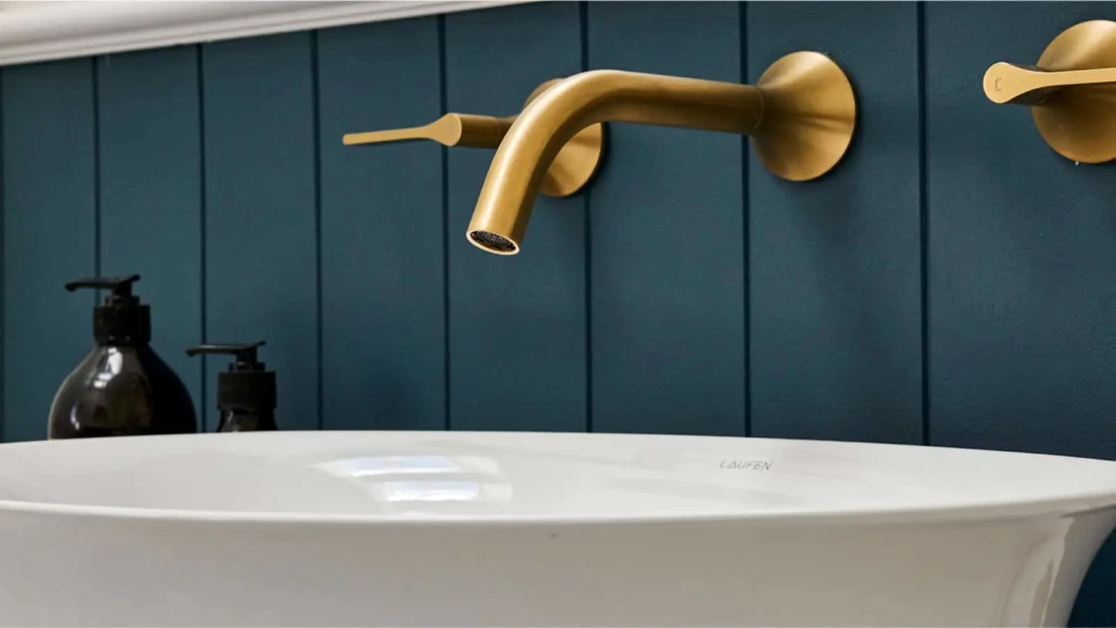 Gold wall-mounted faucet above white Laufen sink with dark teal paneled wall background and black soap dispensers.