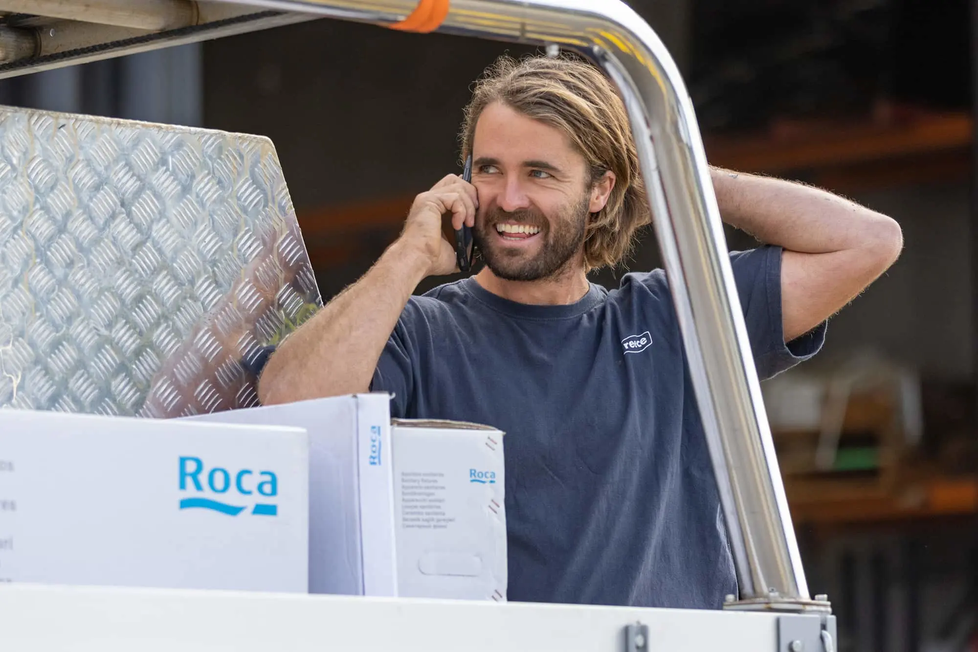 Person talking on phone in work vehicle with Roca boxes visible, wearing blue t-shirt with shoulder-length hair.