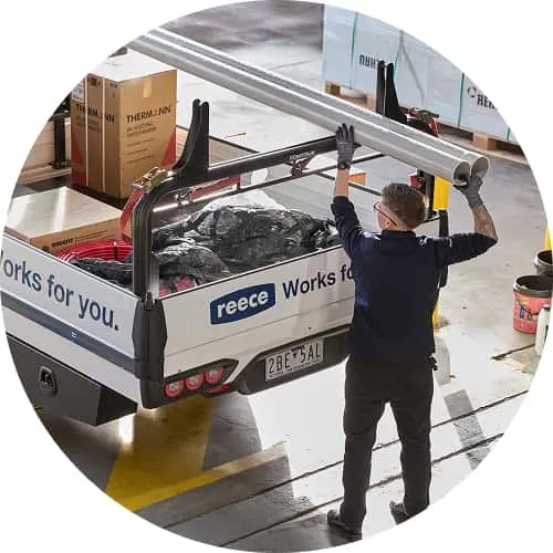 Worker loading metal pipes onto a Reece service truck filled with construction materials in a warehouse.
