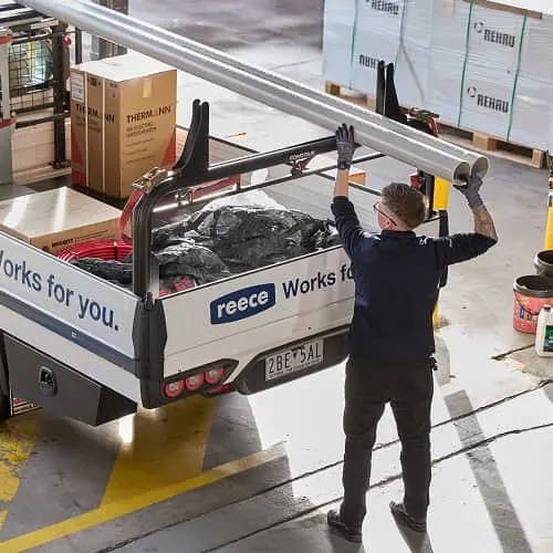 Worker loading metal pipes onto a Reece service truck filled with construction materials in a warehouse.