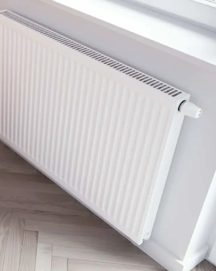 White panel radiator mounted on a wall above light wooden flooring, with temperature control valve visible.