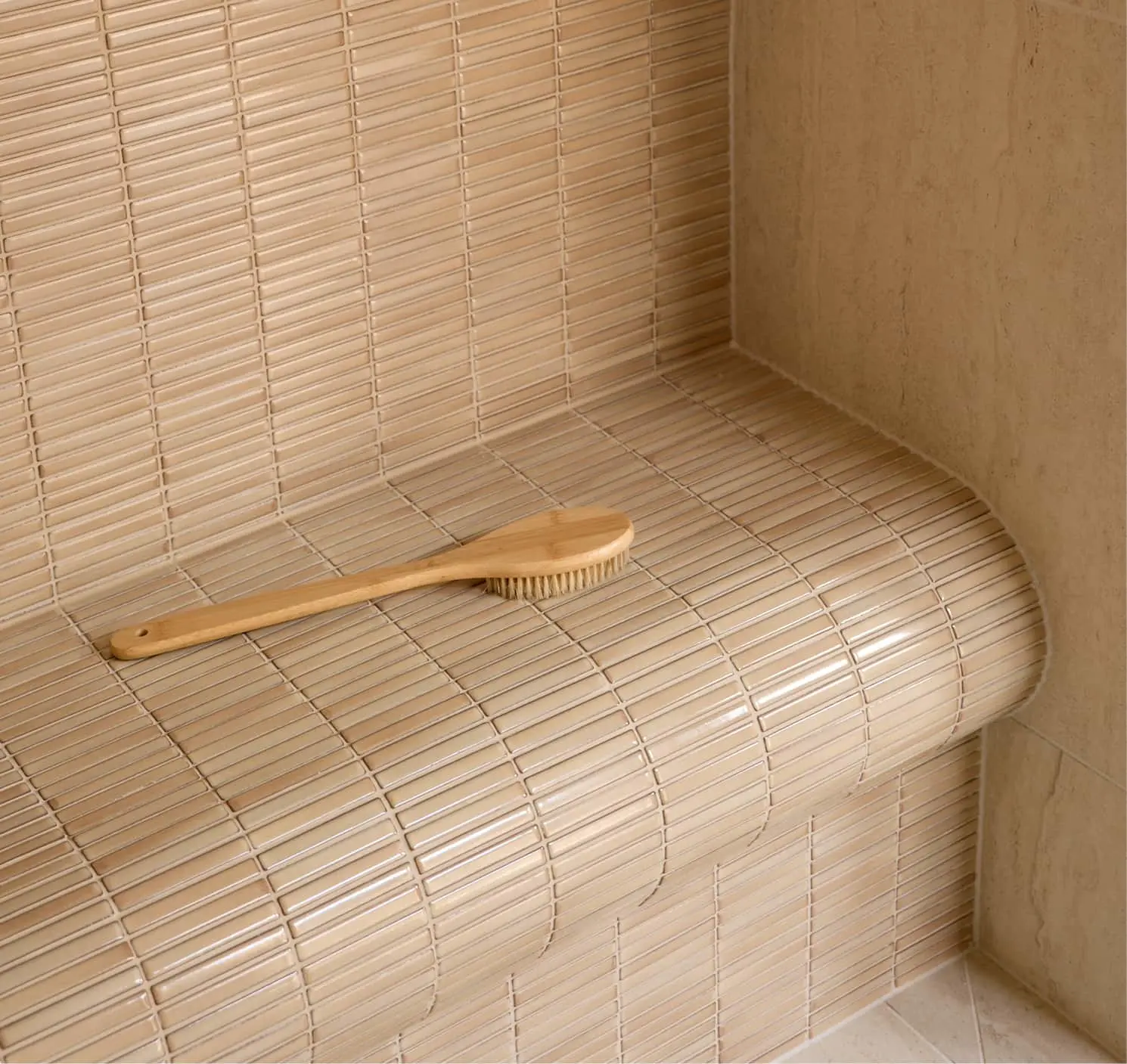 A wooden bath brush resting on a beige mosaic-tiled shower bench with rounded edges.