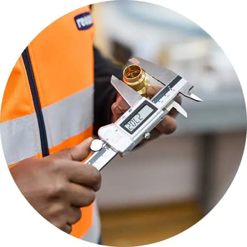 Worker in orange safety vest measuring a brass fitting with digital calipers.