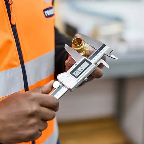 Worker in orange safety vest measuring a brass fitting with digital calipers.
