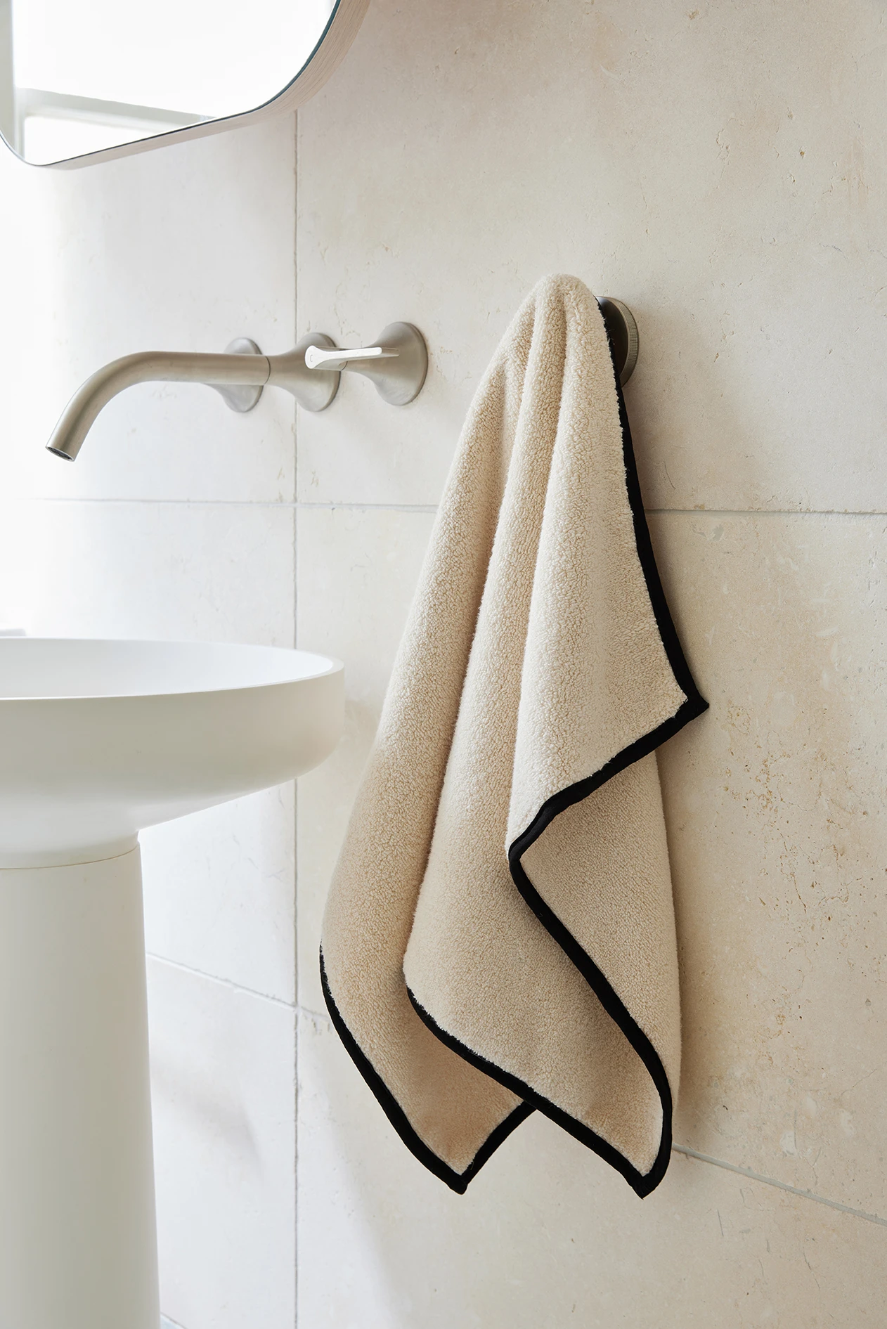A beige hand towel with black trim hangs on a wall hook in a bathroom with stone tiles and a wall-mounted faucet.