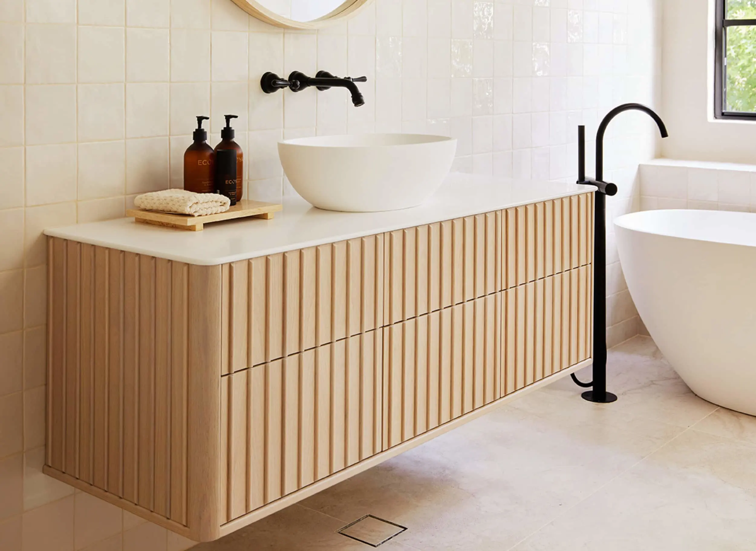 Modern bathroom with wooden vanity, white vessel sink, black fixtures, and freestanding bathtub against cream tiled walls.