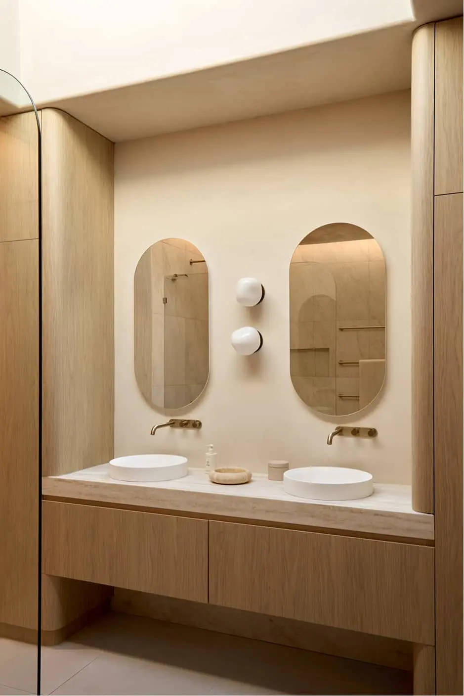 Bathroom vanity with two white vessel sinks, brass wall faucets, oval mirrors, and light wood cabinetry in neutral tones.