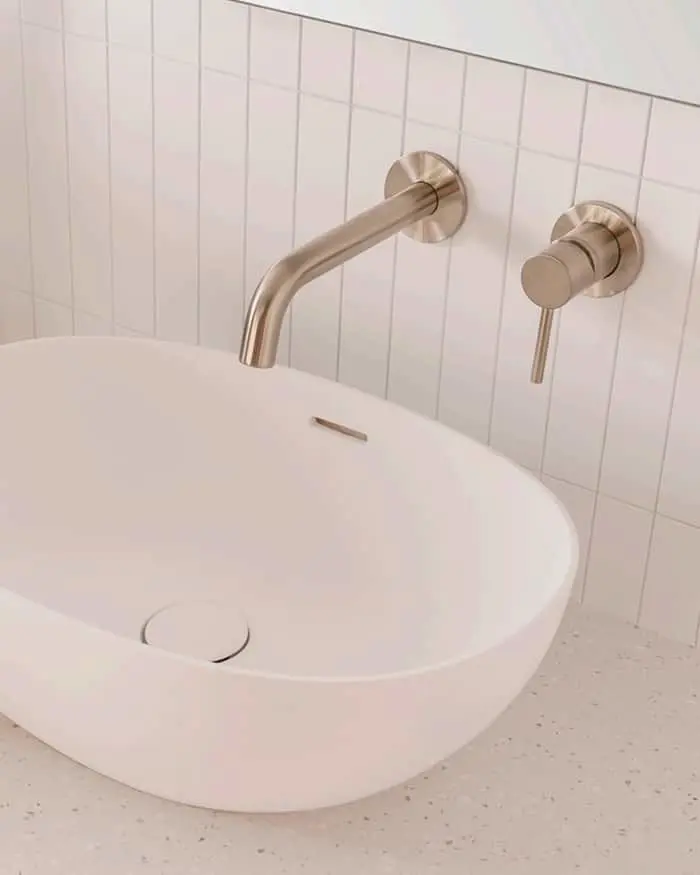 Modern white bathroom sink with brushed gold wall-mounted faucet and handle against white tiled wall.