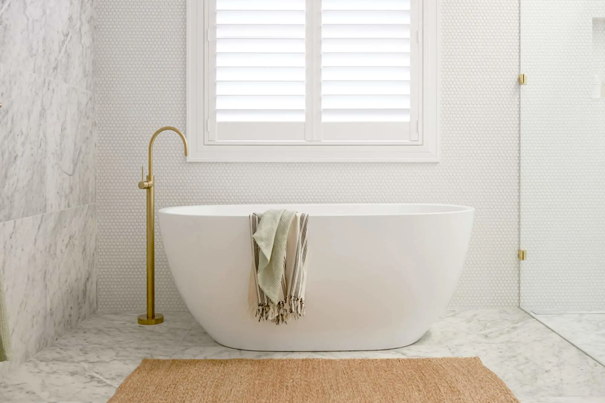 Modern white freestanding bathtub with gold faucet in marble bathroom, featuring window shutters and woven bath mat.