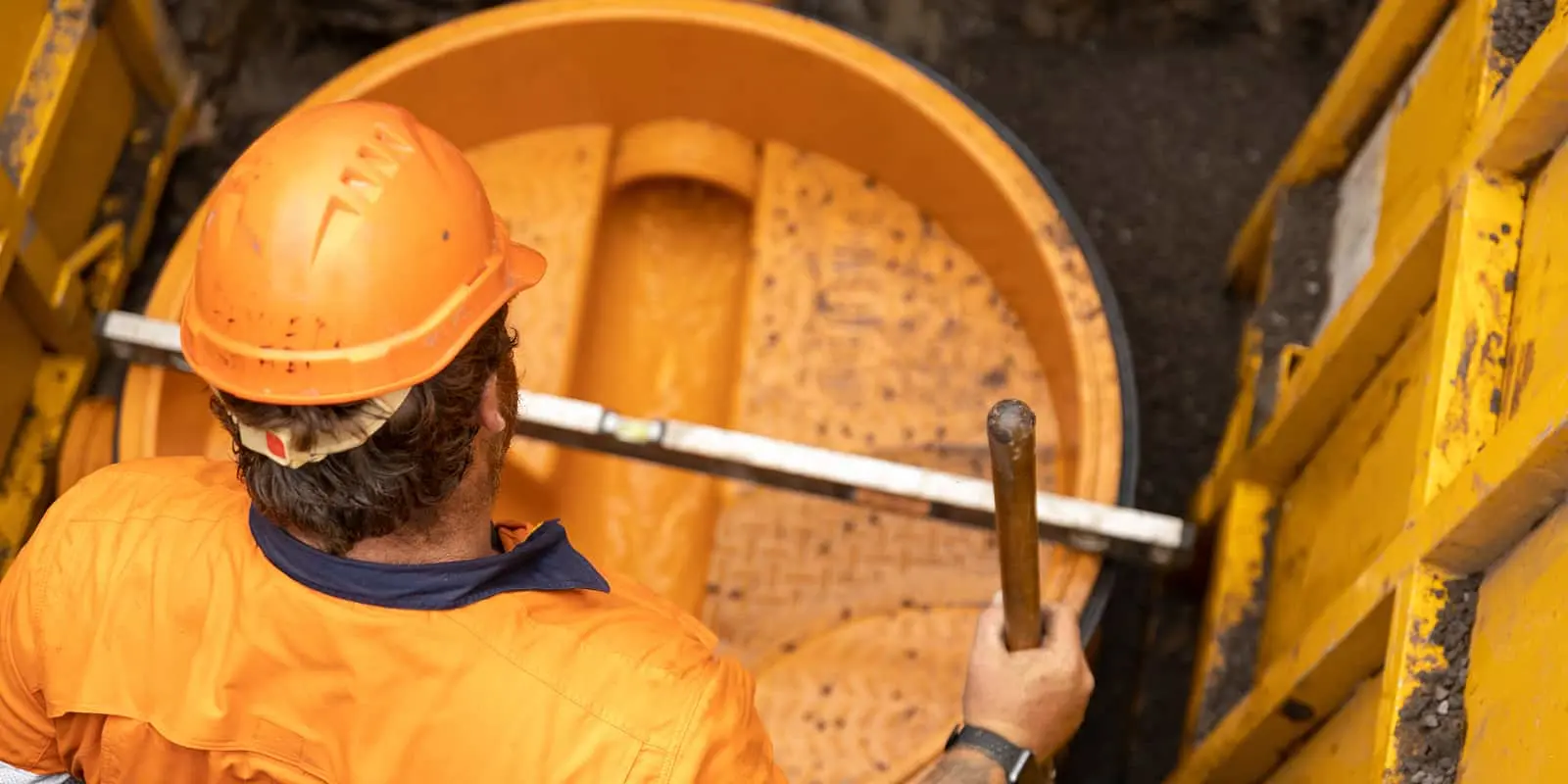 Construction worker in orange hard hat and uniform looking down into a large circular manhole or sewer access point.