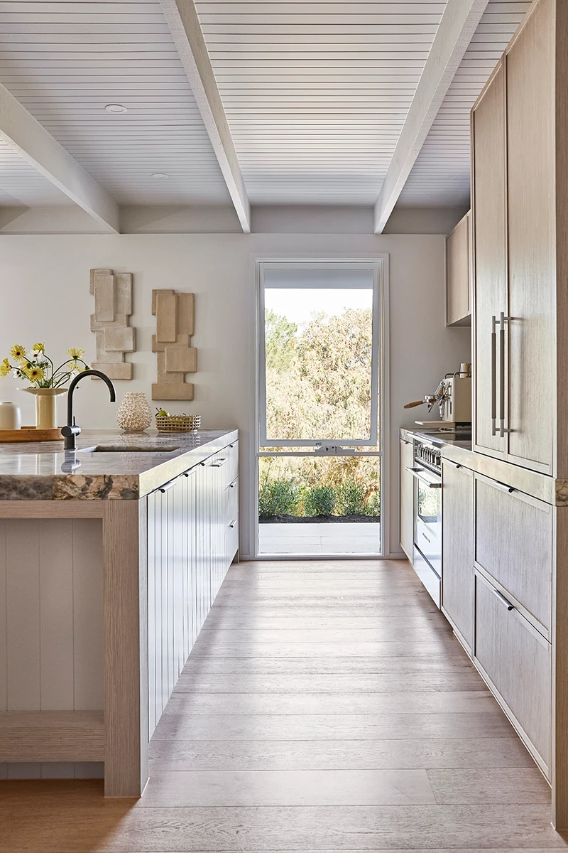 Modern kitchen with light wood cabinets, marble countertop, and glass door overlooking greenery under a white beamed ceiling.