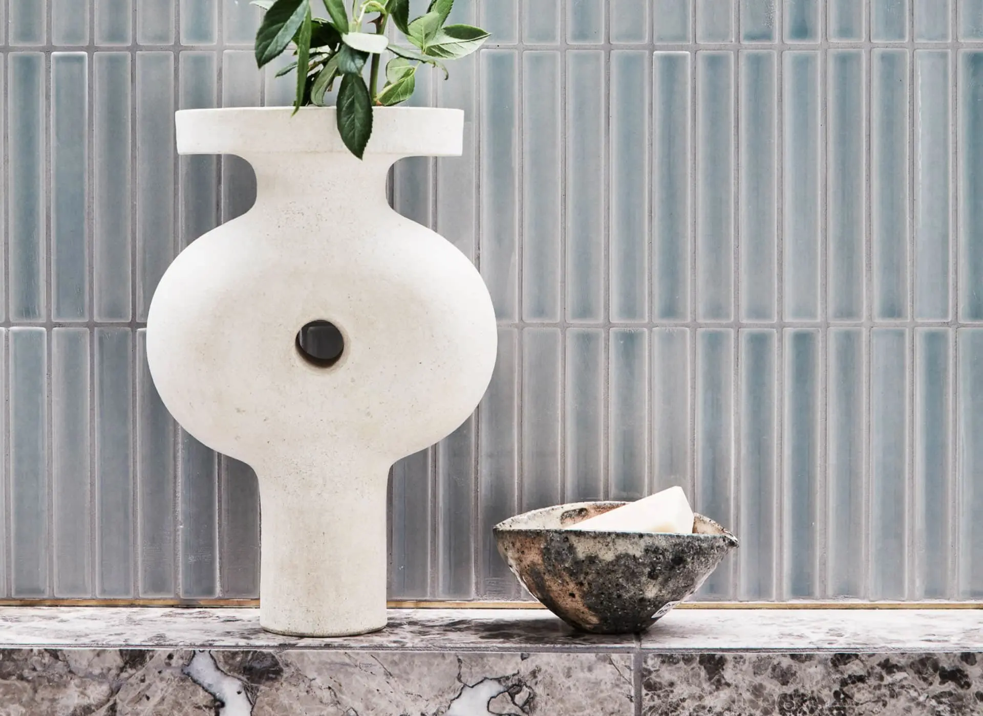 White ceramic vase with green plant next to small textured bowl on marble surface against blue-gray tiled wall.