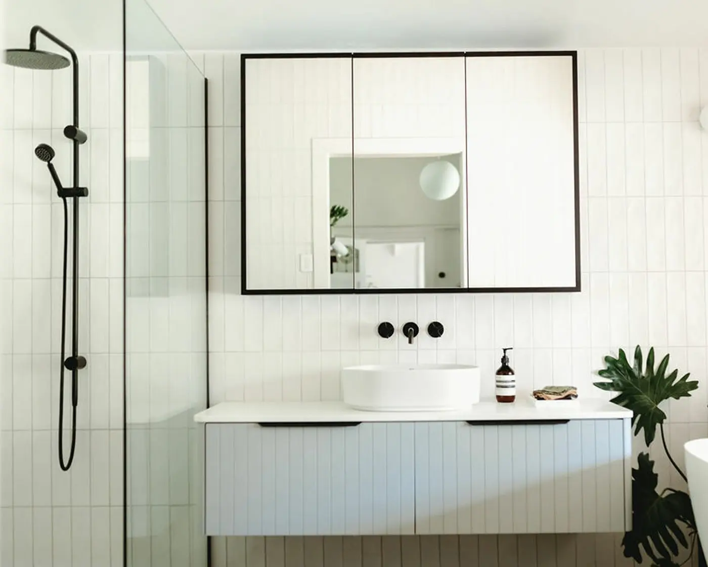 Modern white bathroom with black-framed mirror cabinet, vessel sink, black fixtures, and green plant in corner.