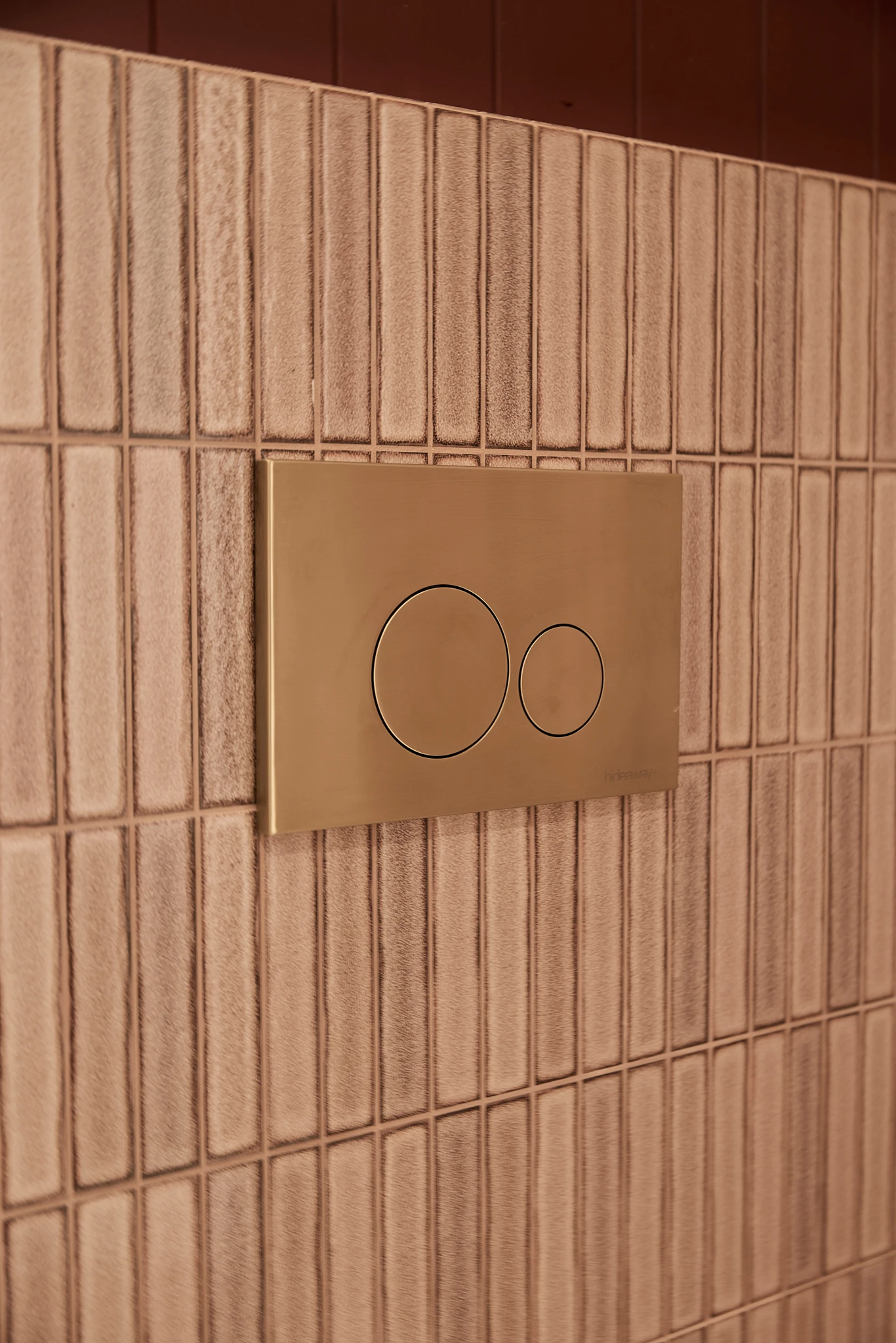 Gold dual-button toilet flush plate mounted on beige vertical tiled bathroom wall.