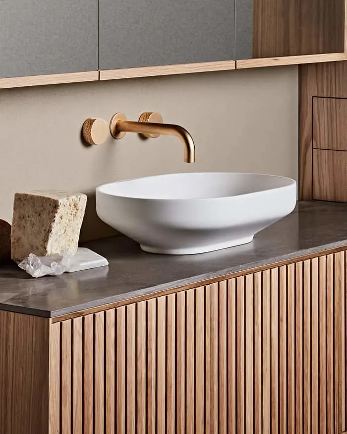 Modern bathroom vanity with white vessel sink, brass wall-mounted faucet, and wooden cabinetry with vertical slats.