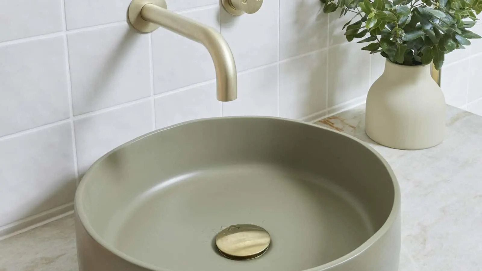 Modern bathroom sink with sage green basin, gold wall-mounted faucet, and small plant in cream vase on marble counter.