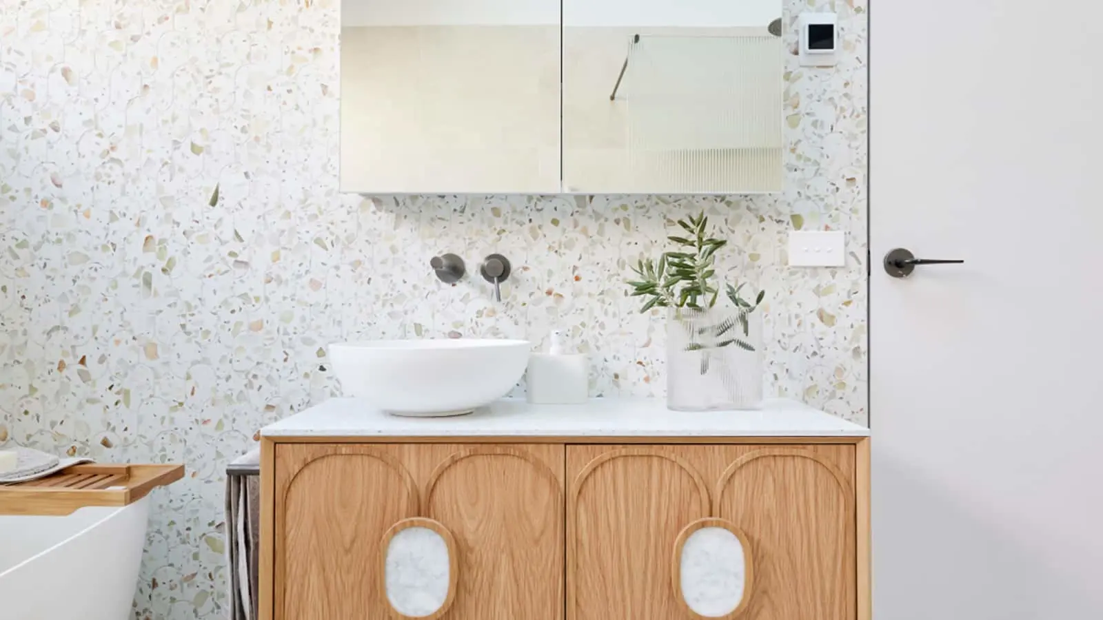 Modern bathroom with terrazzo walls, wooden vanity, white vessel sink, wall-mounted faucet, and small plant in vase.