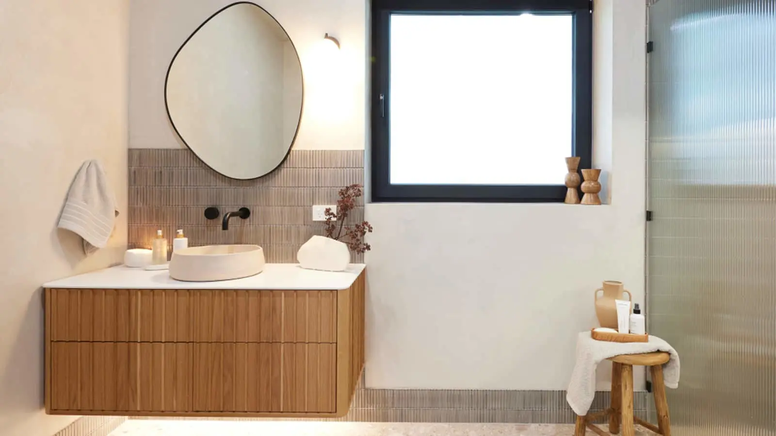 Modern bathroom with wooden vanity, round mirror, vessel sink, black window frame, and wooden stool with toiletries.