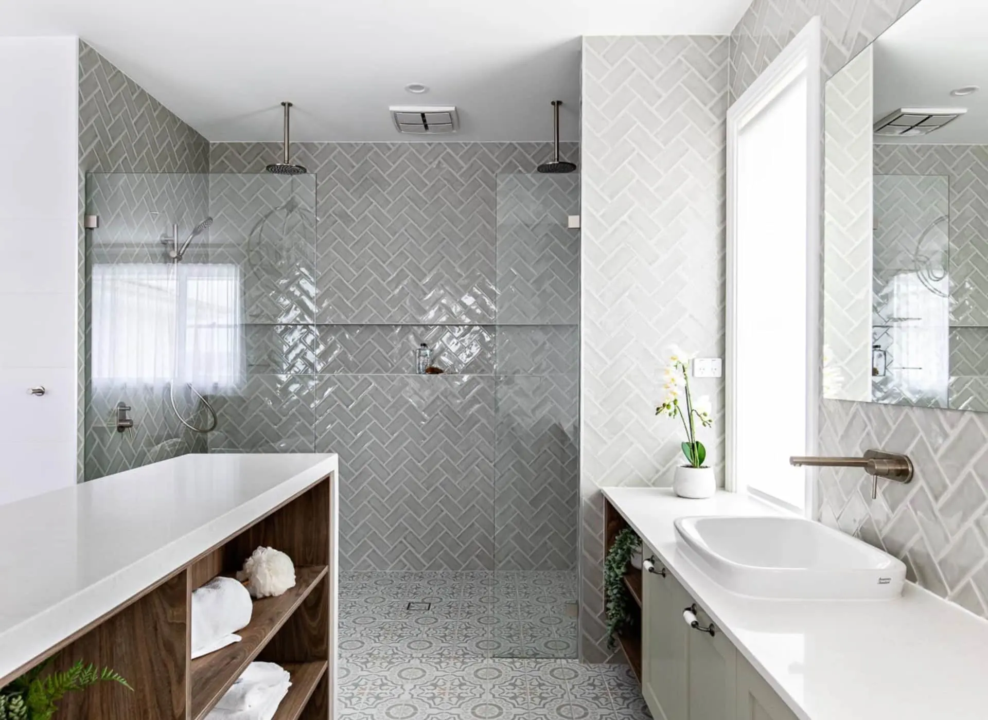 Modern bathroom with gray herringbone tile, glass shower, wooden storage, and white sink vanity.
